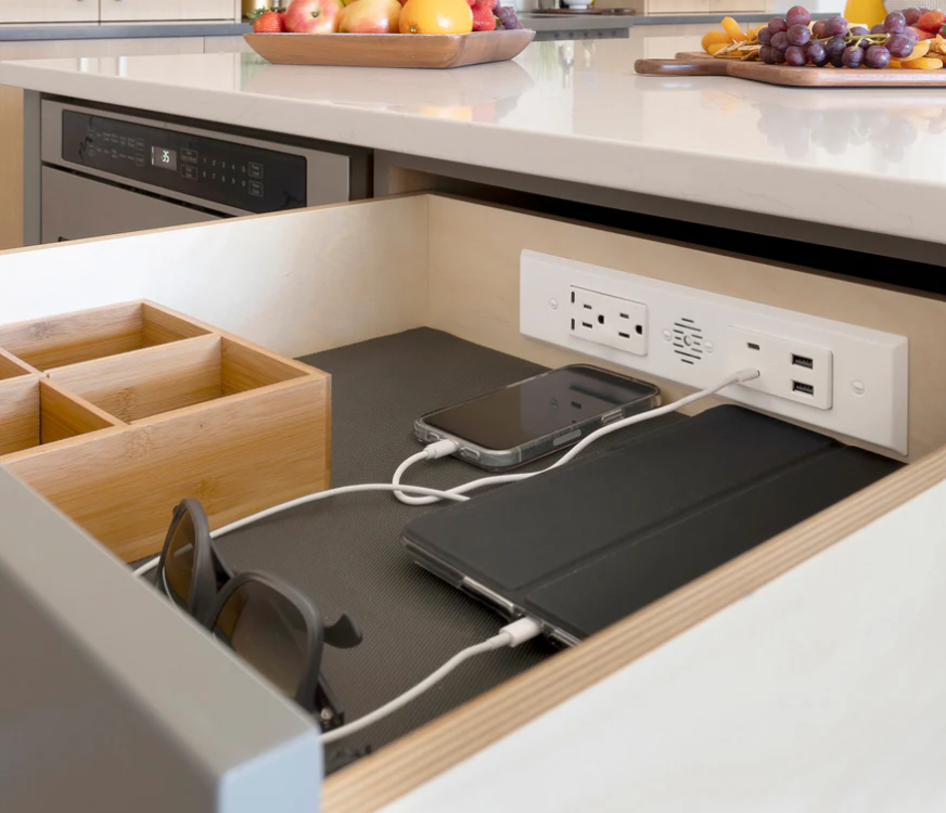 Under cabinet drawer containing built in outlets charging a phone and tablet with a wooden organizer and sunglasses nearby